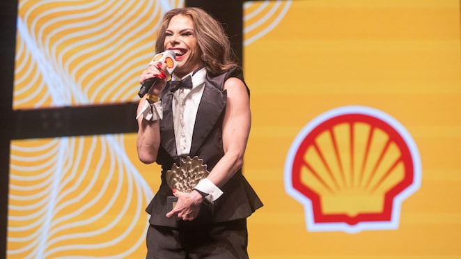Drag queen sorridente segura um troféu em formato de concha dourada enquanto fala ao microfone no palco do Prêmio Shell de Teatro. Ela usa terno preto com camisa branca e gravata borboleta, em frente a um telão com o logotipo da Shell sobre fundo amarelo com listras brancas curvas.