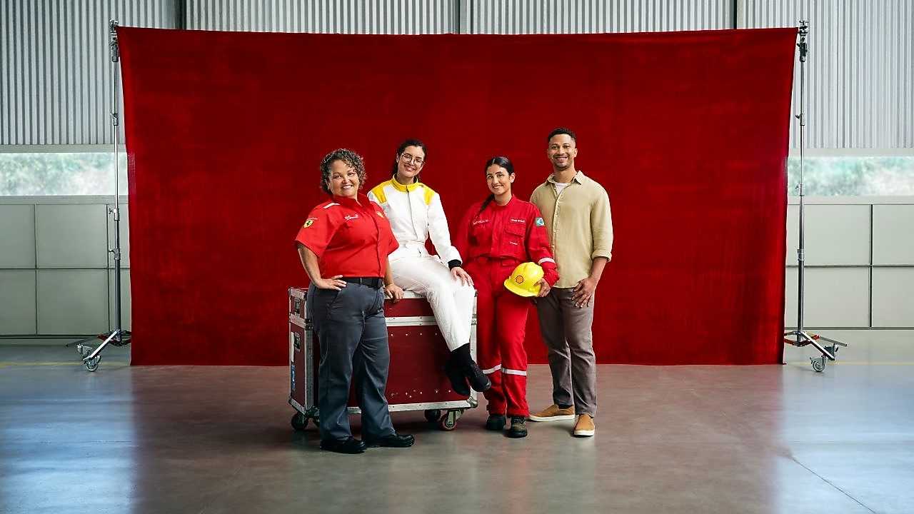Quatro pessoas usando diferentes uniformes profissionais posam em um galpão industrial em frente a um grande painel vermelho. Uma das pessoas está sentada sobre um baú metálico com rodízios, enquanto as outras três permanecem em pé ao redor. Os uniformes incluem roupas em vermelho, branco e bege, e um capacete de segurança amarelo é segurado por uma das pessoas.