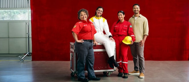 Quatro pessoas usando diferentes uniformes profissionais posam em um galpão industrial em frente a um grande painel vermelho. Uma das pessoas está sentada sobre um baú metálico com rodízios, enquanto as outras três permanecem em pé ao redor. Os uniformes incluem roupas em vermelho, branco e bege, e um capacete de segurança amarelo é segurado por uma das pessoas.
