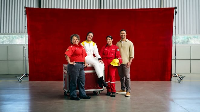 Quatro profissionais posam diante de um fundo vermelho dentro de um galpão industrial. Duas mulheres usam uniformes vermelhos de trabalho, uma pessoa está com macacão branco e outra veste roupa casual; uma delas segura um capacete amarelo. 