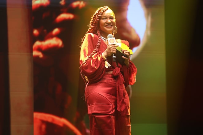 Pessoa em um palco segurando um microfone com a marca Shell e um troféu, sorrindo enquanto fala. Usa um vestido vermelho com mangas volumosas e cabelo trançado longo. Ao fundo, há iluminação em tons quentes e uma projeção ampliada de seu rosto.