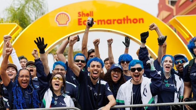 Grupo de jovens participantes da Shell Eco-marathon comemora diante do pórtico amarelo do evento, com braços erguidos e expressões de entusiasmo. Muitos usam uniformes de suas equipes e têm cabelos tingidos de azul.