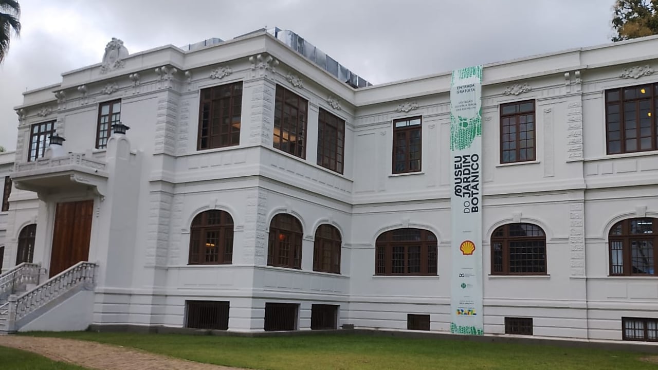 Fachada de um prédio histórico branco do Museu do Jardim Botânico, com janelas de madeira e gramado em frente. Na lateral da fachada, uma faixa vertical exibe o nome do museu e logos de patrocinadores, incluindo o logo da Shell. O céu está nublado.