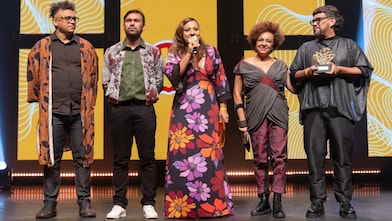 Cinco pessoas estão em um palco iluminado, em frente a um telão amarelo com padrões brancos e pretos. Da esquerda para a direita: um homem de óculos e cabelo grisalho curto usa camisa preta, calça jeans e um kimono marrom estampado; um homem de barba curta veste camisa verde, calça preta e jaqueta estampada; uma mulher no centro segura um microfone e usa vestido longo preto com grandes flores roxas, lilases, vermelhas, laranjas e amarelas; uma mulher com cabelo crespo curto veste blusa cinza transpassada com detalhe bordô, calça roxa e botas pretas; um homem segura um troféu dourado em forma de concha, veste blusa preta larga e calça preta.