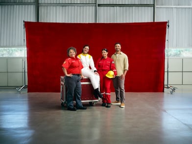 Quatro profissionais posam diante de um fundo vermelho dentro de um galpão industrial. Duas mulheres usam uniformes vermelhos de trabalho, uma pessoa está com macacão branco e outra veste roupa casual; uma delas segura um capacete amarelo.