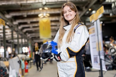 Uma menina sorridente veste um macacão de corrida branco e preto, luvas e segura um capacete azul com viseira transparente. Ela está em um ambiente interno que parece um paddock ou garagem de automobilismo, com placas de identificação amarelas, carros desfocados e outras pessoas ao fundo. O fundo está desfocado, destacando a pessoa em primeiro plano.