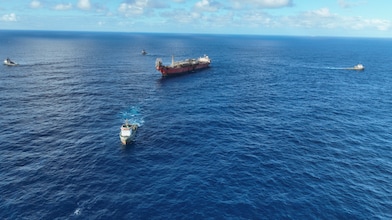 Vista aérea de uma embarcação de grande porte navegando em mar aberto, acompanhada por três embarcações menores ao redor. O oceano azul ocupa a maior parte da imagem, enquanto o horizonte se encontra ao fundo sob um céu claro com nuvens brancas espalhadas. As embarcações deixam rastros na água, destacando o deslocamento em meio à imensidão do mar.