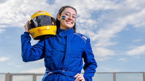 Mulher sorridente veste macacão azul de automobilismo e segura um capacete amarelo da Shell Eco-marathon Brasil sobre o ombro. Ela usa óculos e tem pinturas verdes e amarelas no rosto. Ao fundo, céu azul com nuvens.