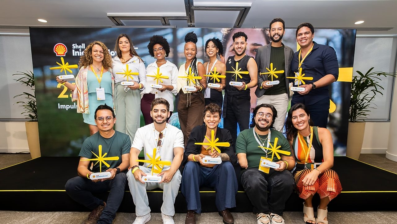 Grupo diverso de pessoas posa para foto em um palco, segurando troféus amarelos em formato de estrela, durante evento da Shell Iniciativa Jovem. Ao fundo, um painel com a marca Shell e elementos visuais do programa.