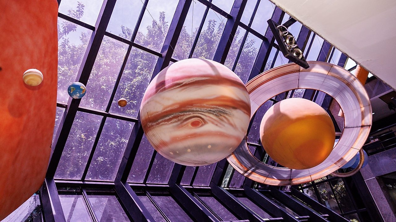 Instalação artística com planetas do sistema solar suspensos no interior de um espaço envidraçado, com destaque para Júpiter e Saturno em primeiro plano, este último com seus anéis visíveis, enquanto outros planetas menores aparecem ao fundo, iluminados pela luz natural que entra pelas grandes janelas.
