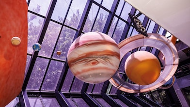 Vista interna de um átrio moderno com teto de vidro, exibindo um modelo suspenso do sistema solar. Grandes esferas detalhadas representam planetas, incluindo um planeta semelhante a Júpiter com faixas visíveis e um planeta com anéis, além de outros menores ao redor. A luz natural entra pelas janelas, e árvores podem ser vistas do lado de fora.