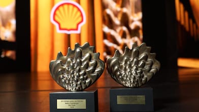 Dois troféus metálicos do 36º Prêmio Shell de Teatro sobre bases pretas, com placas indicando “Melhor Dramaturgia” para São Paulo e Rio de Janeiro (2026), posicionados sobre um palco com iluminação quente; ao fundo, o logotipo da Shell desfocado.