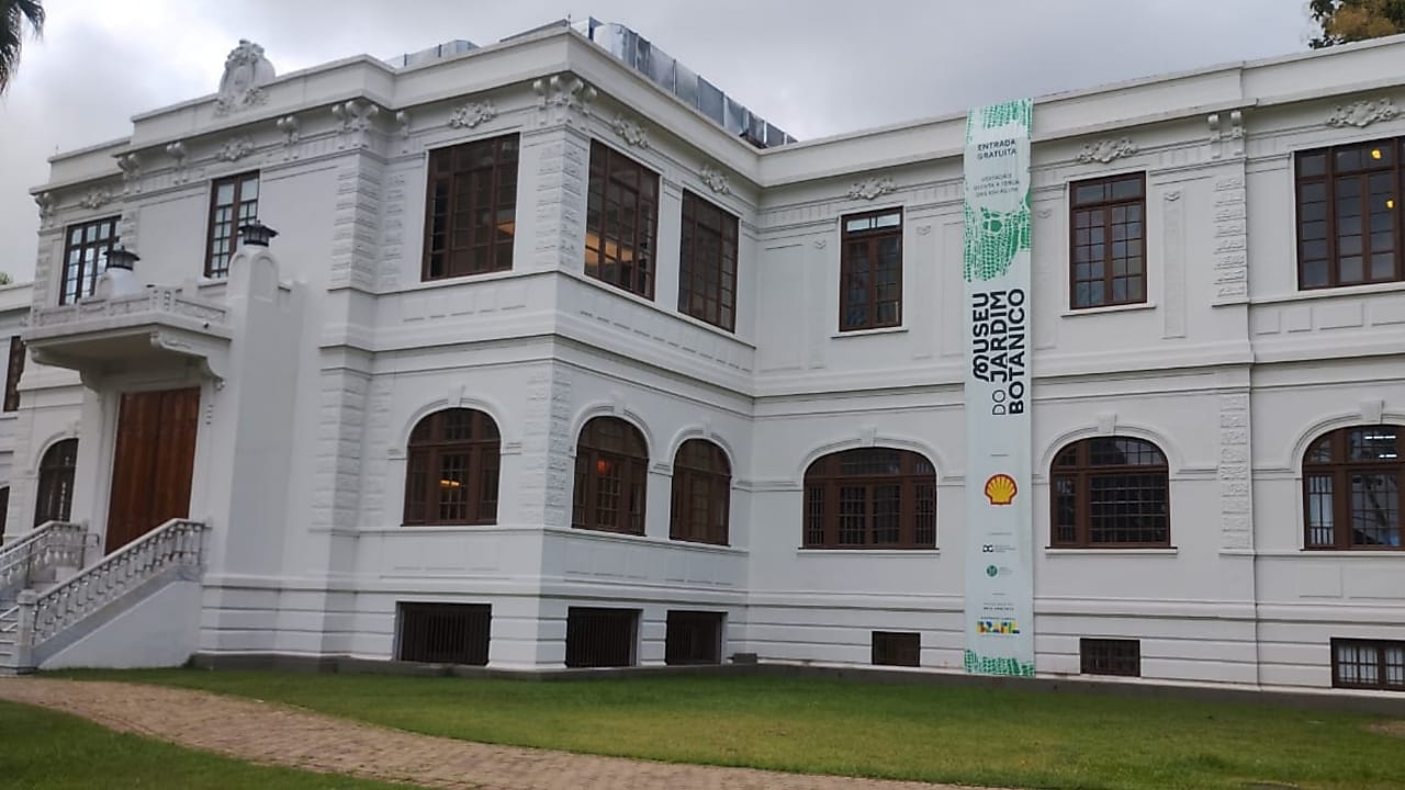 Fachada de um prédio histórico branco do Museu do Jardim Botânico, com janelas de madeira e gramado em frente. Na lateral da fachada, uma faixa vertical exibe o nome do museu e logos de patrocinadores, incluindo o logo da Shell. O céu está nublado.