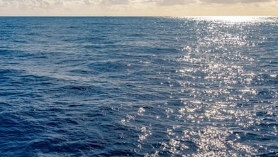 Superfície do oceano sob luz do sol, com pequenas ondas e reflexos dourados brilhando na água azul, sob céu parcialmente nublado.