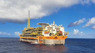 Plataforma flutuante de produção de petróleo (FPSO), de grande porte, com estrutura predominantemente amarela e branca, flutuando em alto-mar sob céu azul com algumas nuvens. A embarcação, equipada com módulos industriais e torre alta, está parcialmente descarregando água pelo casco.