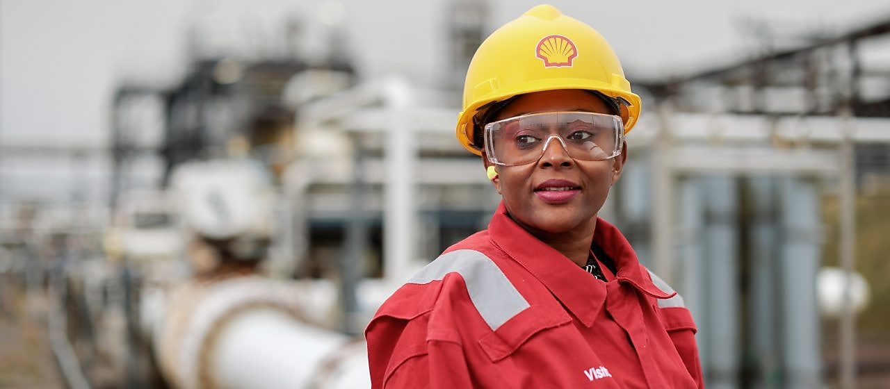 Mulher negra usando capacete amarelo da Shell, óculos de proteção e uniforme vermelho, em ambiente industrial com tubulações e equipamentos ao fundo, olhando de lado.