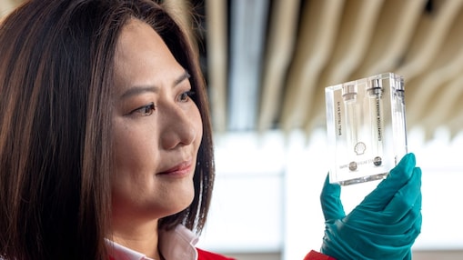 Mulher com jaleco vermelho e luvas de proteção azuis analisa atentamente um pequeno recipiente transparente com amostras em laboratório, segurando-o à altura dos olhos em ambiente técnico e bem iluminado.