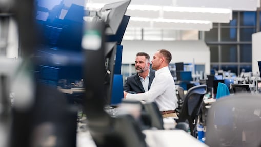 Dois homens conversam em um escritório amplo e moderno, cercados por mesas com computadores e monitores; um deles aponta ou indica algo na tela enquanto o outro observa, em ambiente corporativo com estações de trabalho ao fundo. 