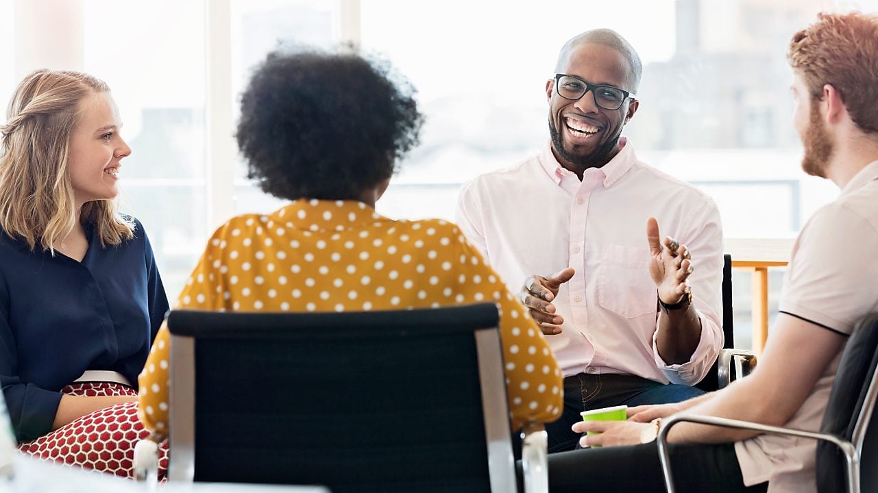 Grupo diverso de pessoas reunidas em um ambiente profissional, com diferentes gêneros, etnias e estilos, sentadas em círculo durante uma conversa colaborativa. A cena transmite inclusão e pluralidade, com participação equilibrada e escuta ativa entre os integrantes, reforçando a ideia de troca de perspectivas e convivência em um espaço de trabalho diverso.