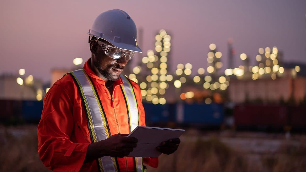 Homem usando capacete, óculos de proteção e uniforme laranja com colete refletivo consulta um tablet em área industrial ao entardecer, com luzes de instalações ao fundo desfocadas.