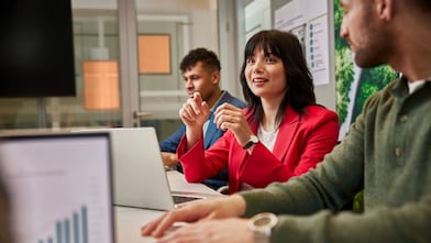 Mulher de blazer vermelho fala e gesticula durante uma reunião de trabalho em escritório; ao seu lado, dois colegas participam da conversa, sentados à mesa com laptops abertos, enquanto gráficos e materiais visuais relacionados a dados aparecem no ambiente.