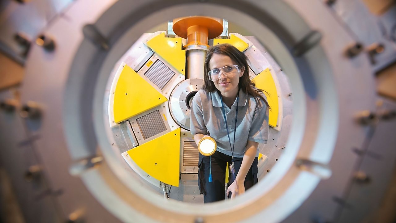 Mulher com óculos de proteção observa o interior de um equipamento industrial circular, segurando uma lanterna; ao redor, estruturas metálicas e painéis amarelos indicam um ambiente técnico e de alta tecnologia.