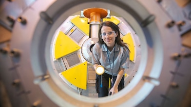 Mulher com óculos de proteção observa o interior de um equipamento industrial circular, segurando uma lanterna; ao redor, estruturas metálicas e painéis amarelos indicam um ambiente técnico e de alta tecnologia. 
