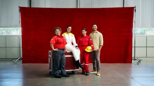 Quatro profissionais posam diante de um fundo vermelho dentro de um galpão industrial. Duas mulheres usam uniformes vermelhos de trabalho, uma pessoa está com macacão branco e outra veste roupa casual; uma delas segura um capacete amarelo. 