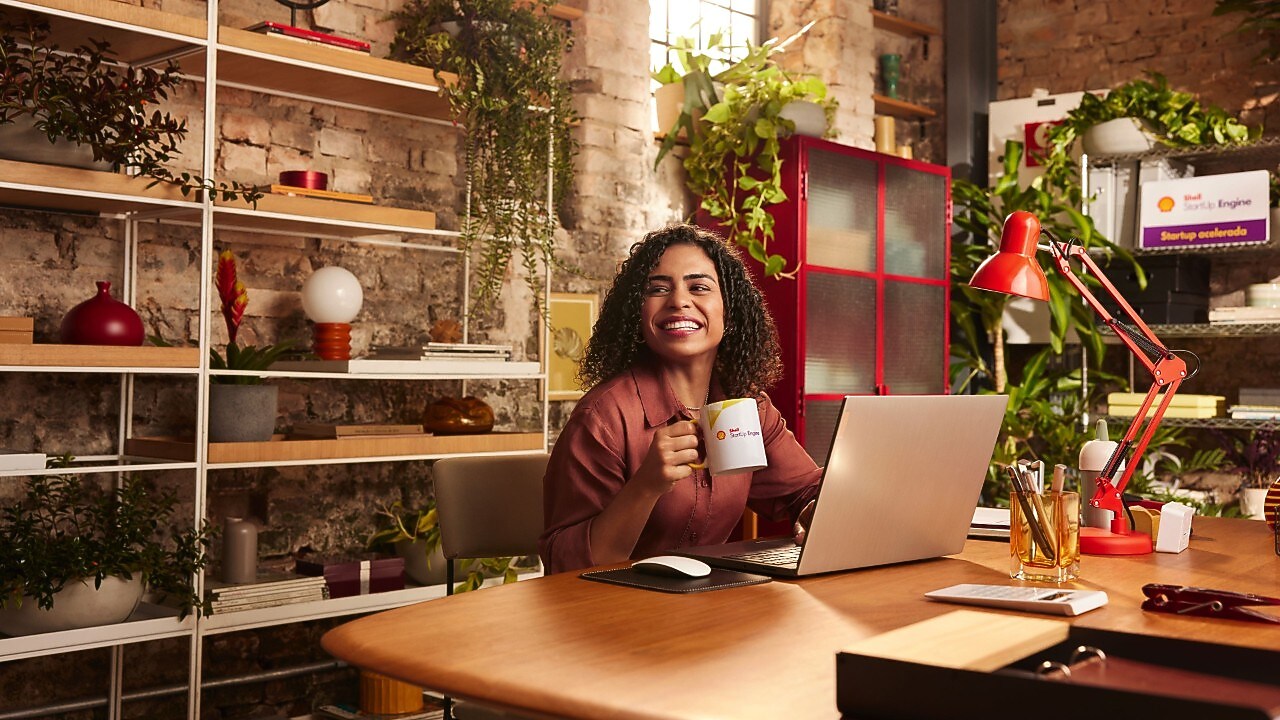 Mulher negra segurando caneca em frente ao seu computador em sala toda decorada com plantas