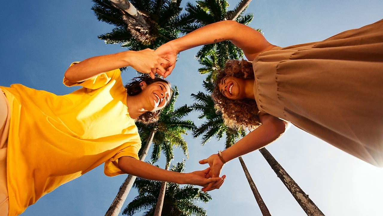 Na imagem, duas mulheres estão de mãos dadas e sorriem, embaixo de palmeiras localizadas no Jardim Botânico do Rio de Janeiro