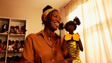 Na imagem, mulher segura uma boneca preta que está vestida com uma roupa de sereia.