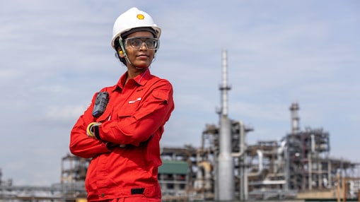 Uma mulher de pele negra olha para o horizonte com os braços cruzados. Ela está vestindo um macacão vermelho e um capacete com a logo de concha da Shell. Ao fundo está uma refinaria.