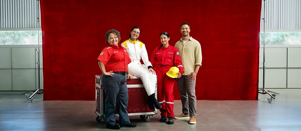 Quatro profissionais posam diante de um fundo vermelho dentro de um galpão industrial. Duas mulheres usam uniformes vermelhos de trabalho, uma pessoa está com macacão branco e outra veste roupa casual; uma delas segura um capacete amarelo.