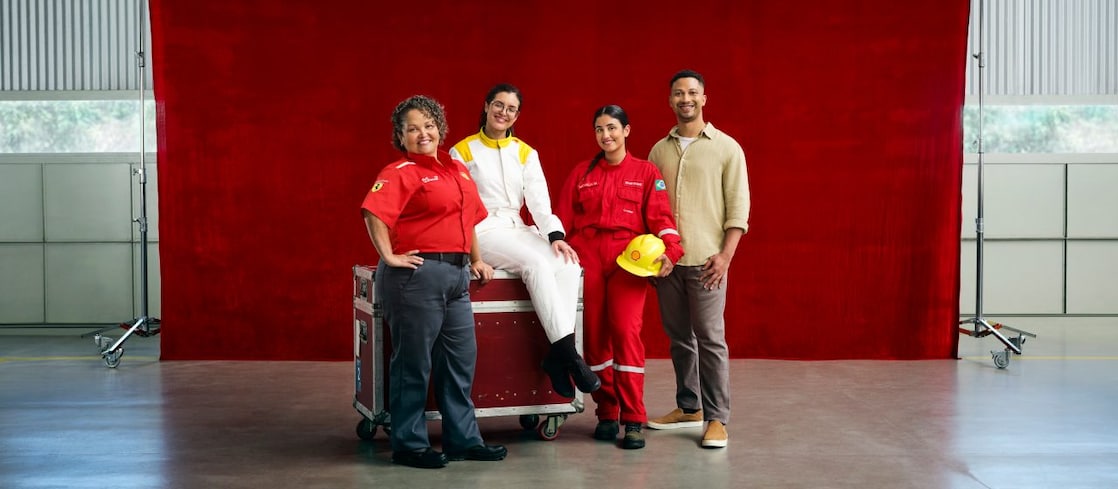 Quatro profissionais posam diante de um fundo vermelho dentro de um galpão industrial. Duas mulheres usam uniformes vermelhos de trabalho, uma pessoa está com macacão branco e outra veste roupa casual; uma delas segura um capacete amarelo. 