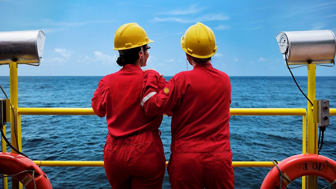 Dois profissionais com macacões vermelhos e capacetes amarelos observam o mar a partir de uma plataforma offshore, apoiados em uma grade amarela, com equipamentos de segurança ao redor.