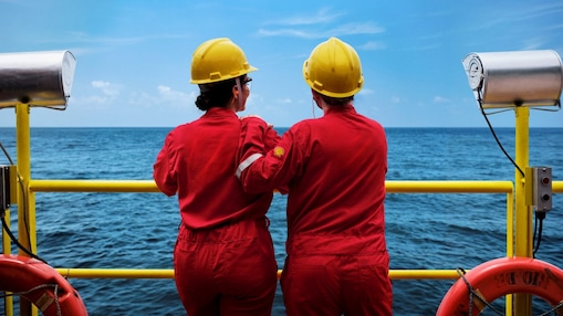 Dois profissionais com macacões vermelhos e capacetes amarelos observam o mar a partir de uma plataforma offshore, apoiados em uma grade amarela, com equipamentos de segurança ao redor.
