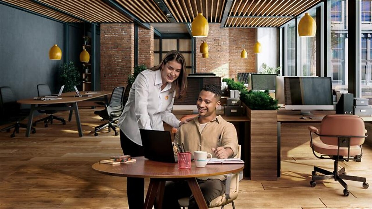 Pessoa em pé orienta um colega sentado enquanto ambos analisam um notebook sobre uma mesa em um escritório moderno, com iluminação quente, plantas e estações de trabalho ao fundo.