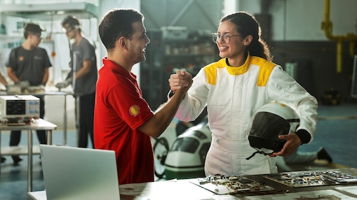 Homem com camisa vermelha da Shell e mulher com macacão branco segurando um capacete apertam as mãos em um laboratório, com equipamentos eletrônicos e outras pessoas trabalhando ao fundo.