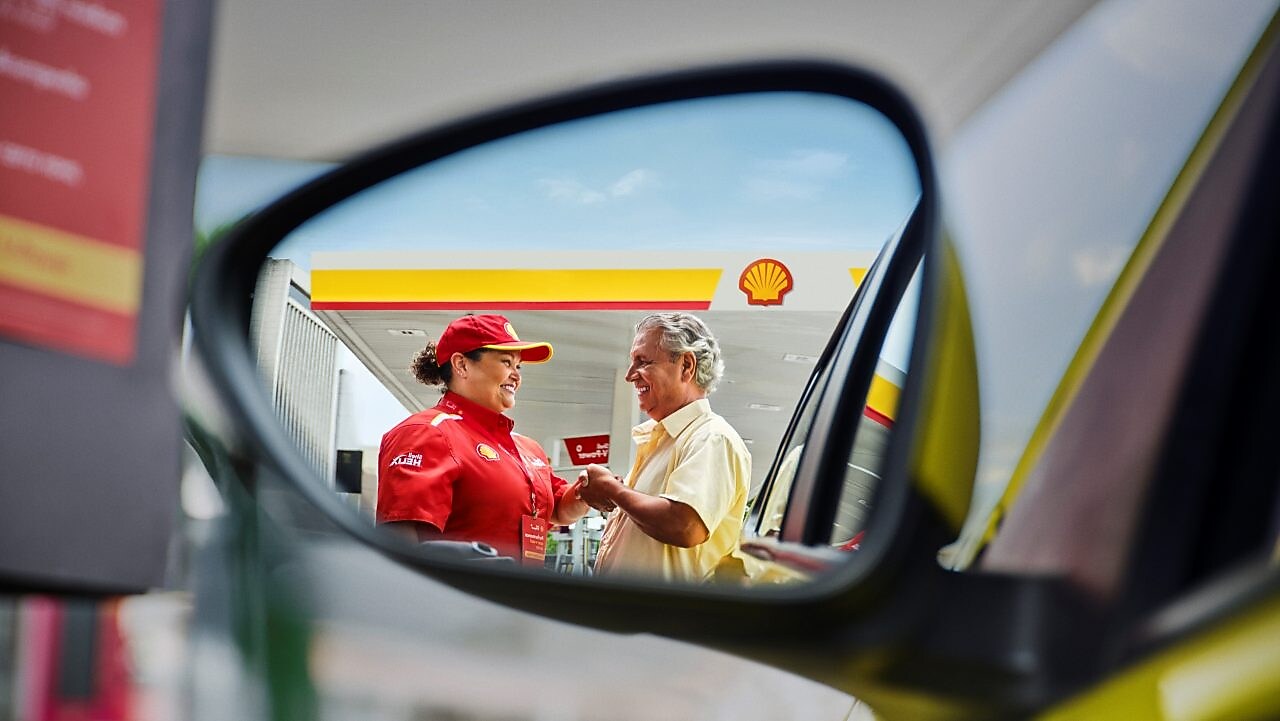 Funcionária de posto Shell, vestindo uniforme vermelho e boné, conversa e sorri para um cliente ao lado do carro, cena vista pelo reflexo do retrovisor.