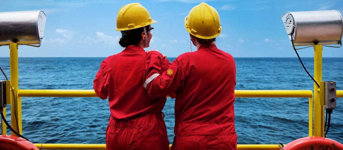 Duas pessoas com macacões vermelhos com logo da Shell e capacetes amarelos observam o mar a partir de uma plataforma offshore, apoiadas em um corrimão amarelo, com equipamentos de segurança ao redor. 