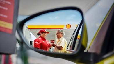 Funcionária de posto Shell, vestindo uniforme vermelho e boné, conversa e sorri para um cliente ao lado do carro, cena vista pelo reflexo do retrovisor.