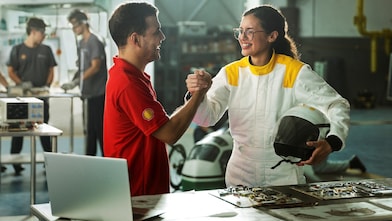 Homem com camisa vermelha da Shell e mulher com macacão branco segurando um capacete apertam as mãos em um laboratório, com equipamentos eletrônicos e outras pessoas trabalhando ao fundo.