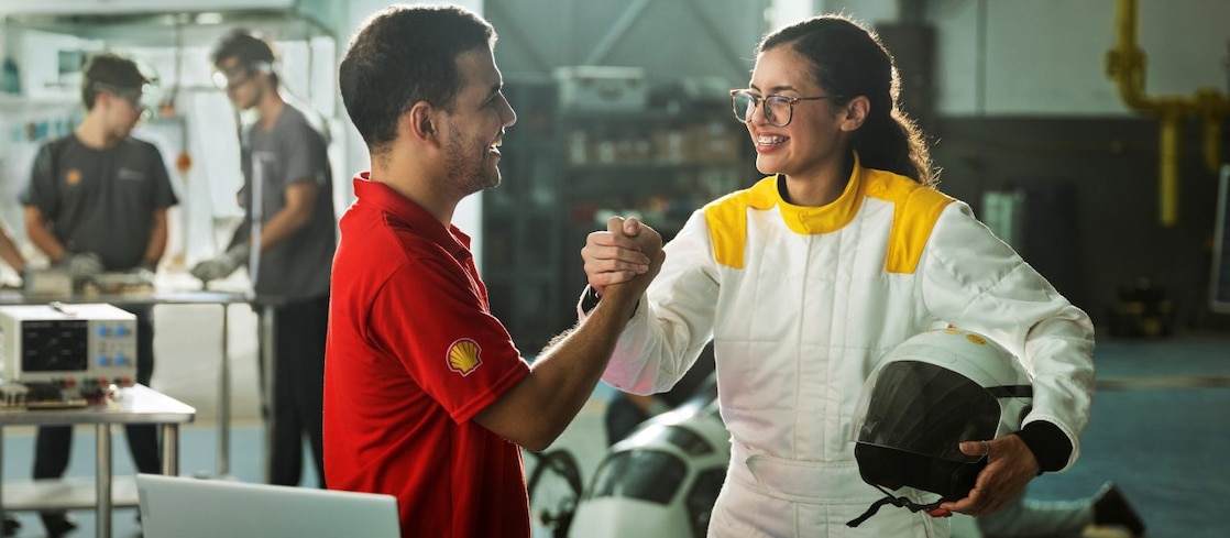 Duas pessoas em um laboratório apertam as mãos em sinal de parceria: um homem com camisa vermelha com o logo da Shell e uma jovem com macacão branco segurando um capacete, com equipamentos eletrônicos e colegas trabalhando ao fundo.