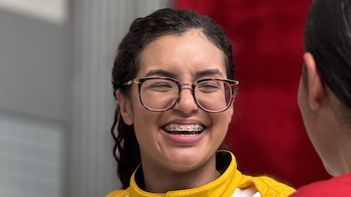 Menina jovem em primeiro plano usando óculos de armação escura e aparelho ortodôntico nos dentes, vestindo um macacão de piloto amarelo. A pessoa está de frente para outra pessoa parcialmente visível à direita da imagem, e está sorrindo para essa pessoa. O fundo da imagem está desfocado.