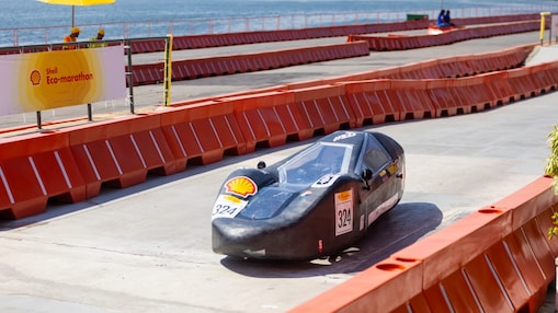 Carro experimental compacto e aerodinâmico, de cor preta e formato alongado, identificado com o número 324 e o logotipo da Shell, percorre uma pista cercada por barreiras laranjas durante o Shell Eco-marathon. Ao fundo, há a Baía de Guanabara, com o Museu do Amanhã visível, além de guarda-sóis amarelos e algumas pessoas observando o trajeto em um dia ensolarado.