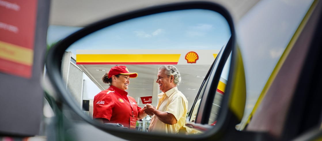 Reflexo no espelho lateral de um carro mostrando uma frentista uniformizada da Shell sorrindo e cumprimentando um cliente idoso em um posto de combustíveis. Ao fundo, vê‑se a cobertura do posto com o logotipo da Shell.