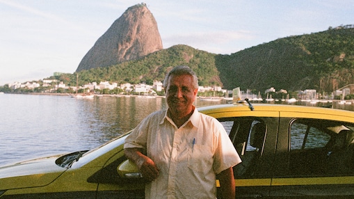 Um homem sorridente está parado ao lado de um carro amarelo à beira da água, com o Pão de Açúcar aparecendo ao fundo. A cena ocorre durante o dia, com água calma e prédios visíveis na orla.
