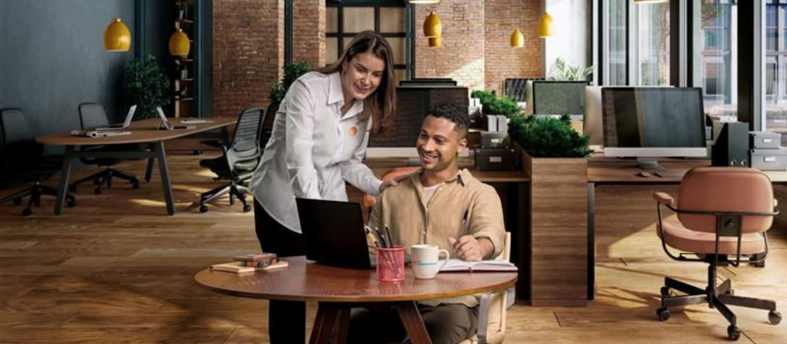 Uma mulher em pé orienta um homem sentado enquanto ele usa um notebook em um ambiente de escritório moderno, com mesas, cadeiras e luminárias ao fundo.