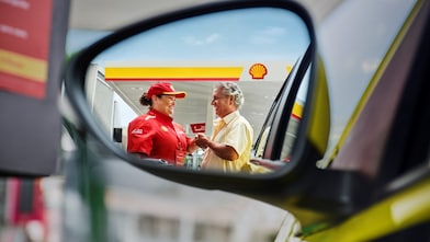 Funcionária de posto Shell, vestindo uniforme vermelho e boné, conversa e sorri para um cliente ao lado do carro, cena vista pelo reflexo do retrovisor.