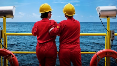 Duas pessoas com macacões vermelhos e capacetes amarelos observam o mar a partir de uma plataforma offshore, apoiadas em um corrimão amarelo, com equipamentos de segurança ao redor.